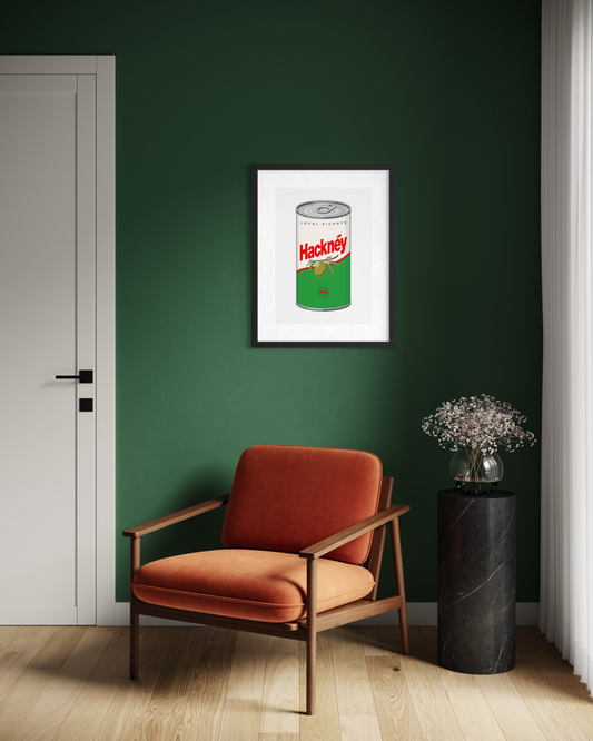 A framed art print of a vintage Hackney olive can poster hangs on a dark green wall in a modern room setting with an orange armchair and a black marble side table.