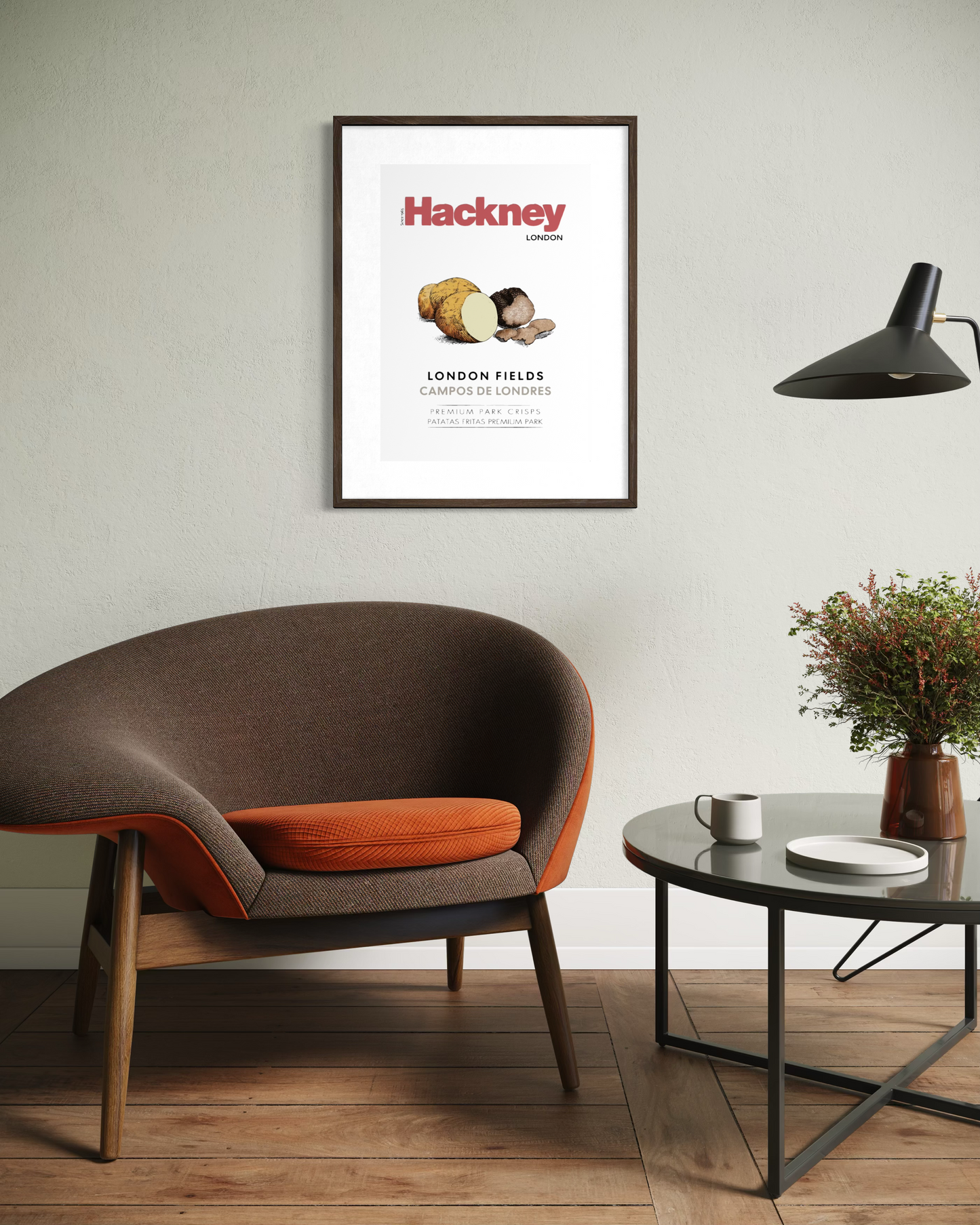 A framed art print featuring a Hackney London poster is displayed on a light gray wall in a modern living room setting with a brown and orange armchair, a black side table, and a lamp.