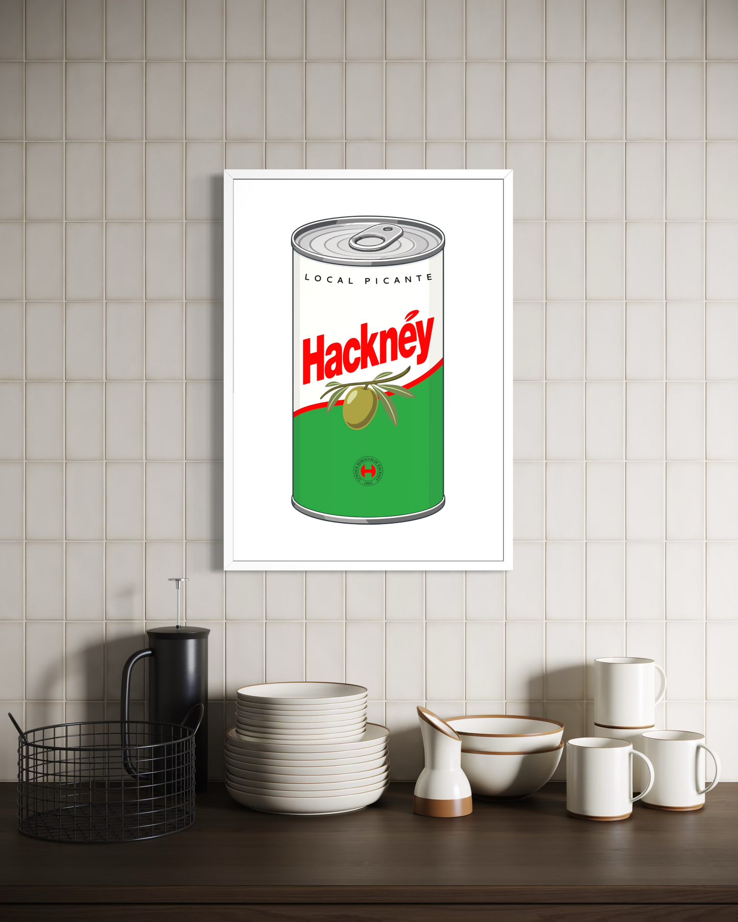 A framed wall art print featuring a vintage-style canned olives design with the word 'Hacknéy' in red, displayed on a beige tiled wall above a kitchen counter with white dishes and a black basket.