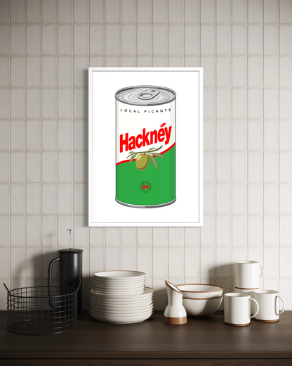 A framed wall art print featuring a vintage-style canned olives design with the word 'Hacknéy' in red, displayed on a beige tiled wall above a kitchen counter with white dishes and a black basket.