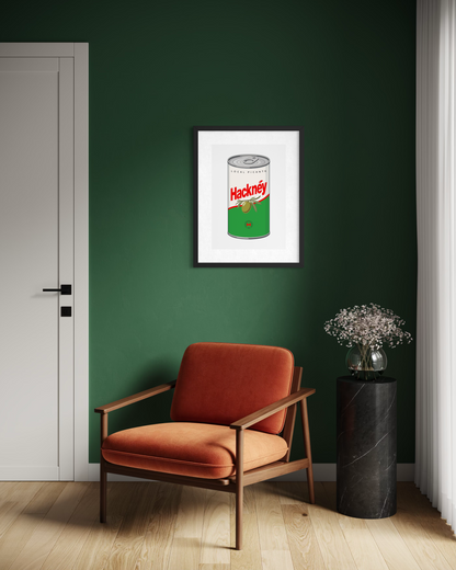 A framed art print of a vintage Hackney olive can poster hangs on a dark green wall in a modern room setting with an orange armchair and a black marble side table.