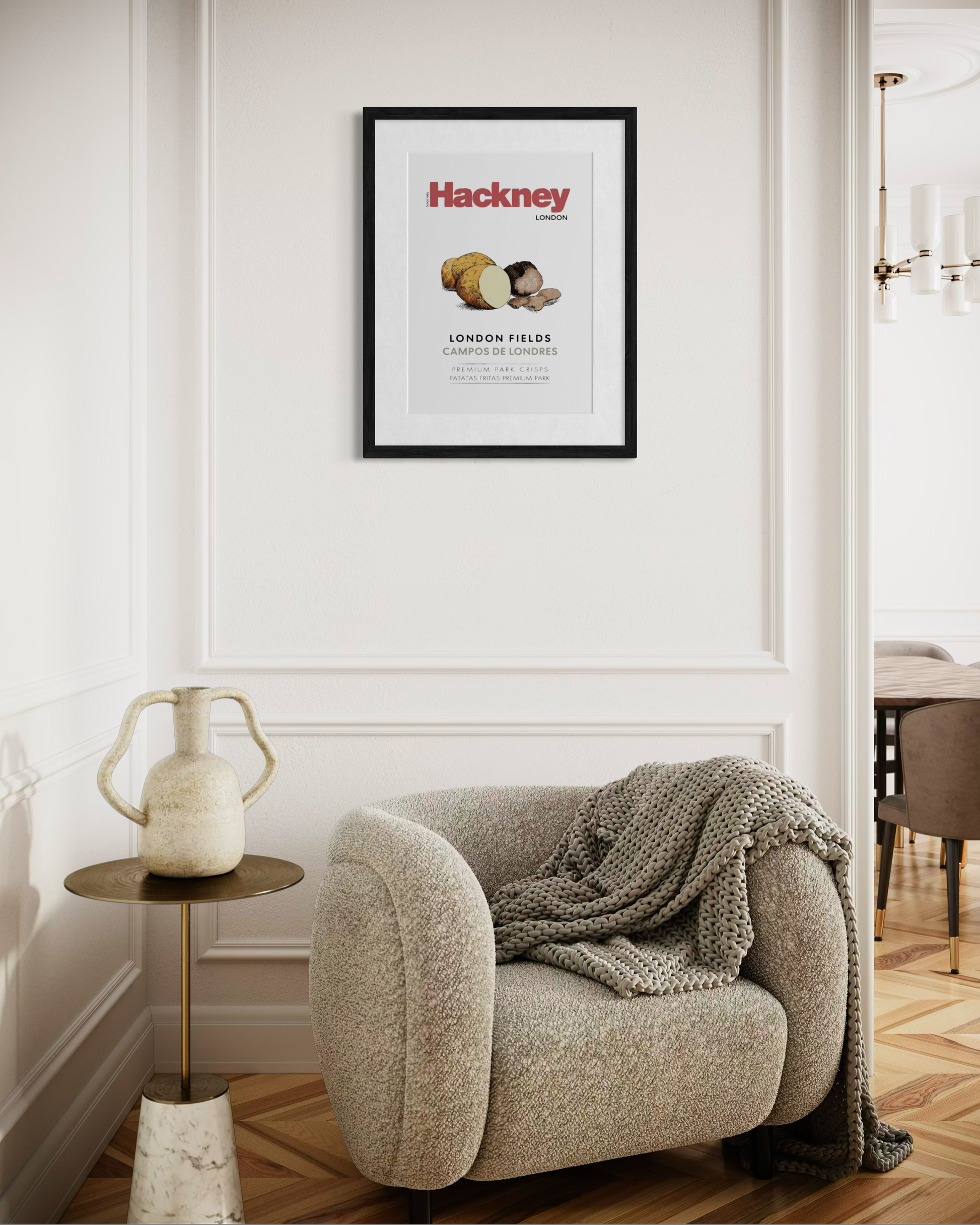 A framed Hackney poster hangs on a white wall above a beige armchair with a chunky knit throw blanket in a styled living room setting.