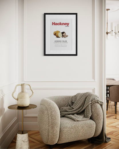 A framed Hackney poster hangs on a white wall above a beige armchair with a chunky knit throw blanket in a styled living room setting.