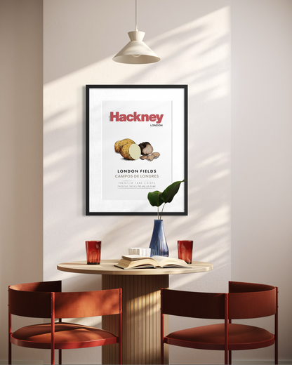 A framed Hackney London poster hangs on a wall above a small round table with two rust-red chairs, a pendant light, and a vase in a sunlit interior.