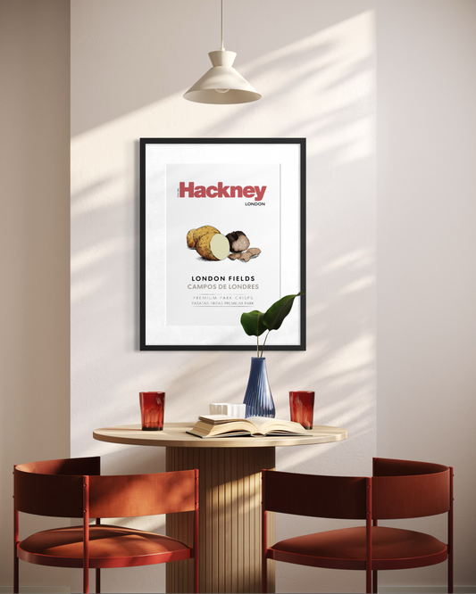 A framed Hackney London poster hangs on a wall above a small round table with two rust-red chairs, a pendant light, and a vase in a sunlit interior.