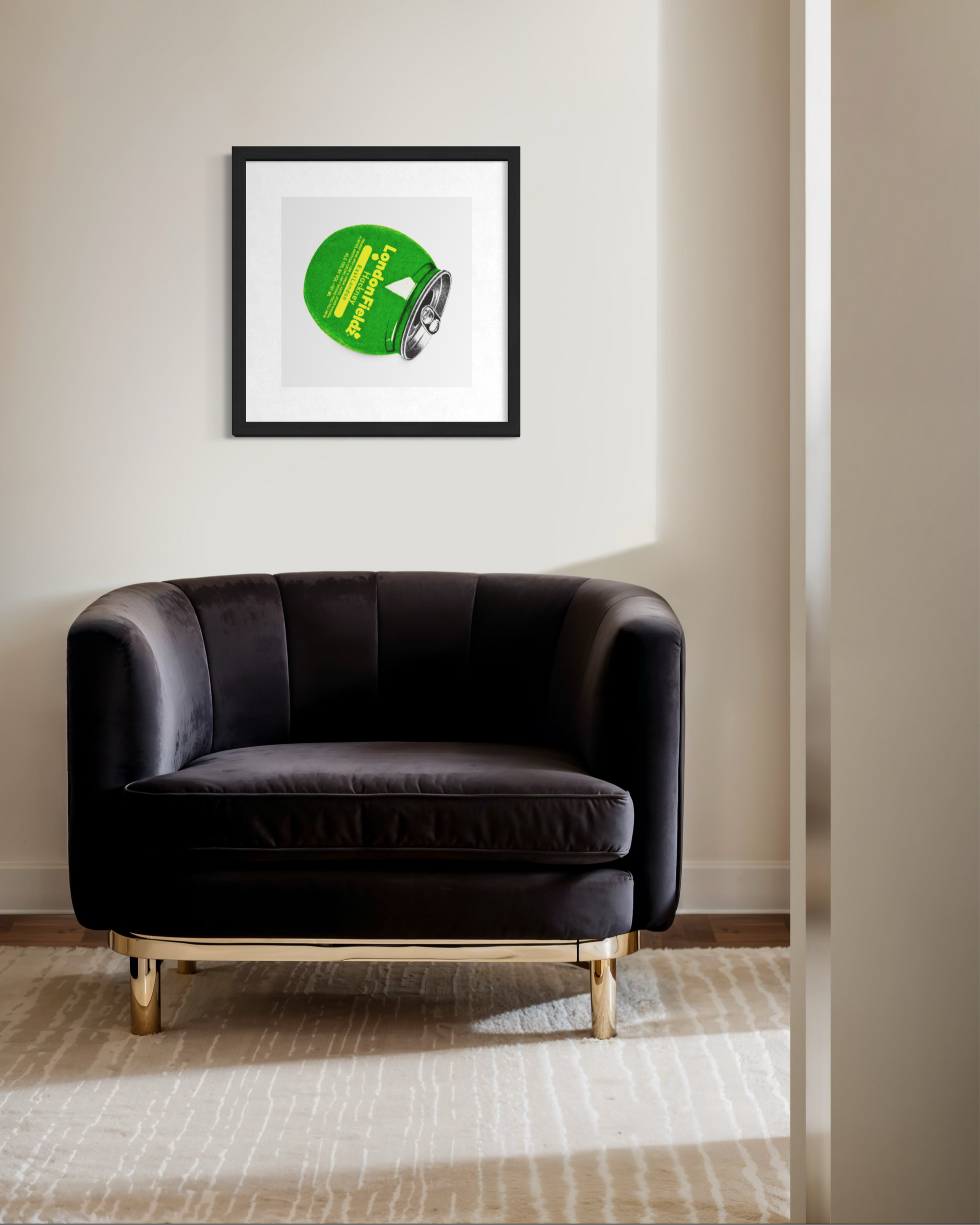 A framed art print featuring a green circular graphic with yellow text and a small black-and-white illustration, displayed on a beige wall above a dark brown velvet armchair with gold legs in a modern room setting.