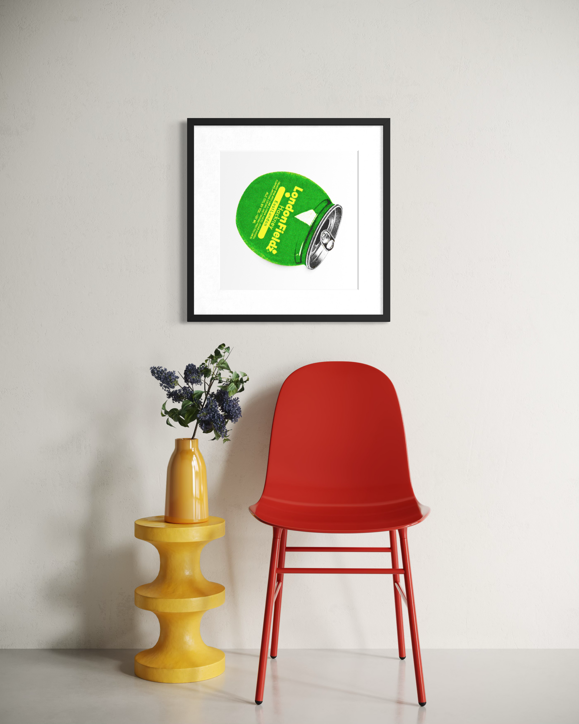 A framed art print featuring a green circular graphic with yellow text, displayed on a light gray wall above a red chair and a yellow side table with flowers.
