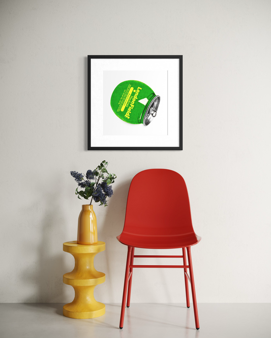 A framed art print featuring a green circular graphic with yellow text, displayed on a light gray wall above a red chair and a yellow side table with flowers.